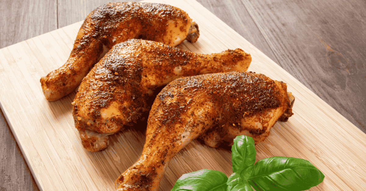 Three seasoned rotisserie chicken legs placed on a wooden cutting board with a fresh basil leaf.