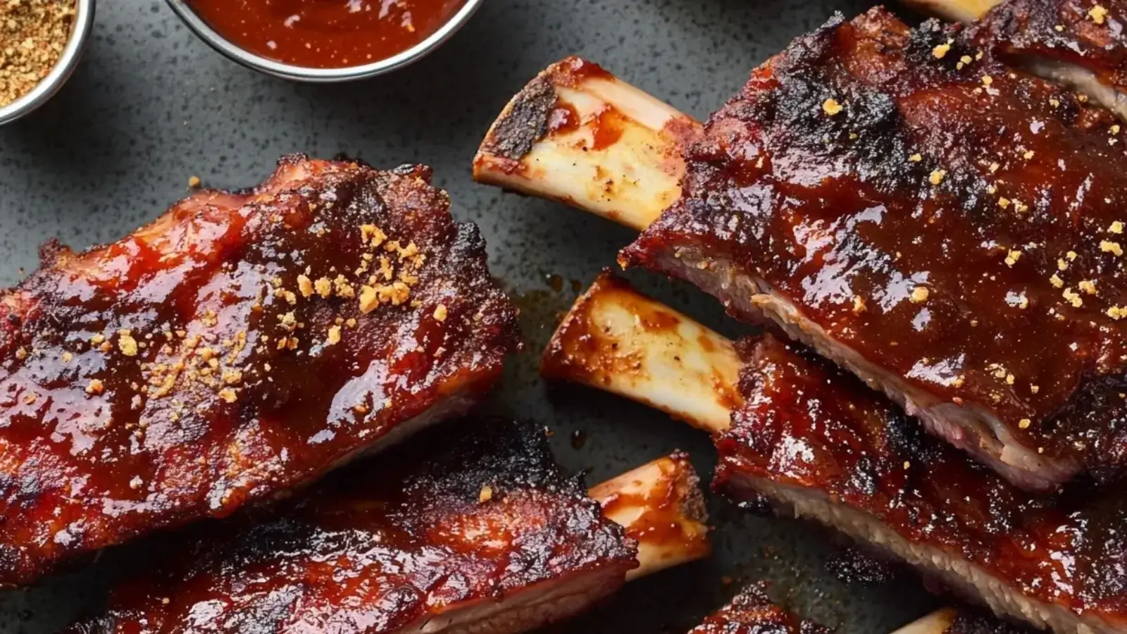 Close-up of juicy beef back ribs with a shiny barbecue glaze, set on a rustic table with sauce and spices.